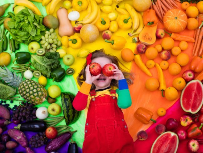 Kleines Mädchen inmitten von farblich sortierten Obst- und Gemüsesorten