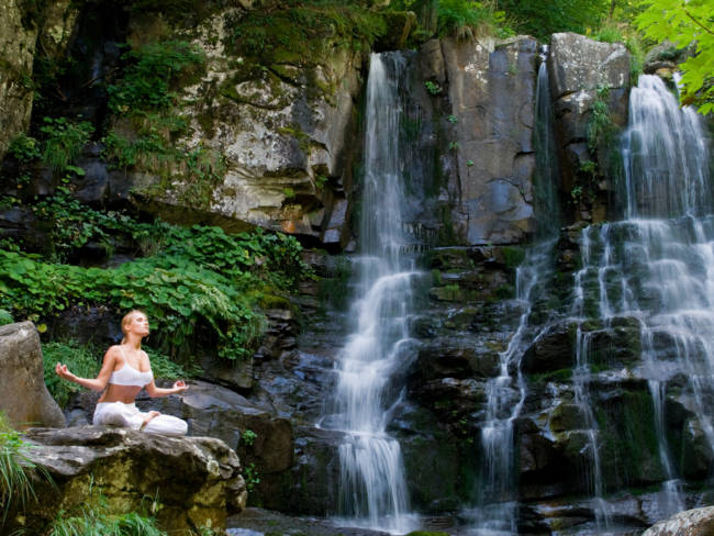 Frau meditiert in freier Natur neben einem Wasserfall.